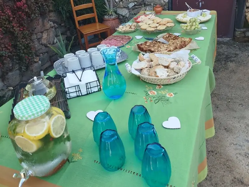 Mesa ao ar livre com pão, queijos, bolos, fruta e água com limão, junto a um muro de pedra
