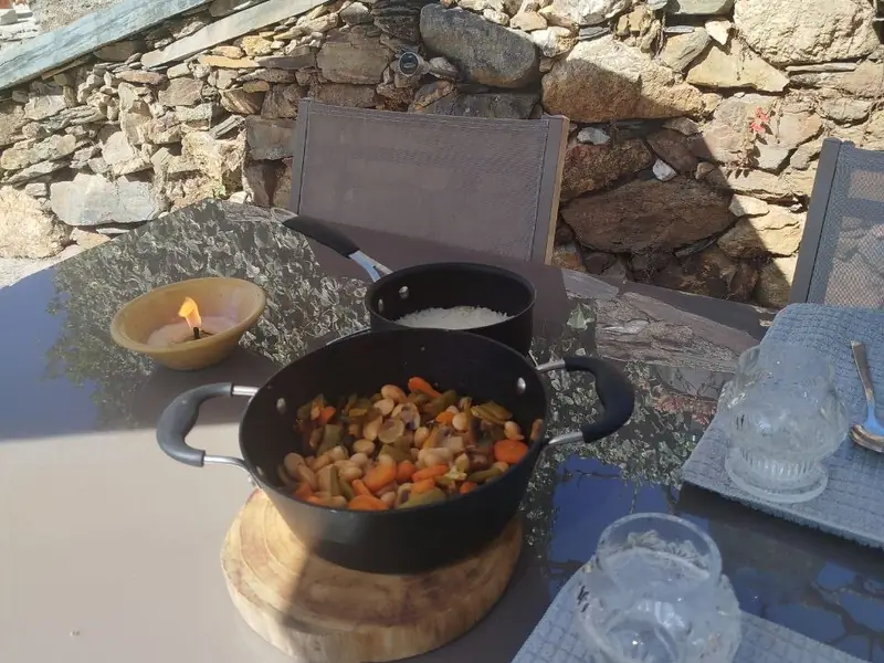 Mesa ao ar livre com um tacho de feijão e legumes, uma panela de arroz e uma vela, junto a um muro de pedra