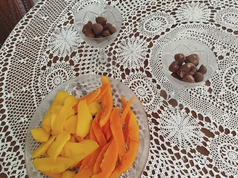 Taça de vidro com fatias de fruta e duas taças com pequenos doces escuros, sobre uma toalha de croché branca